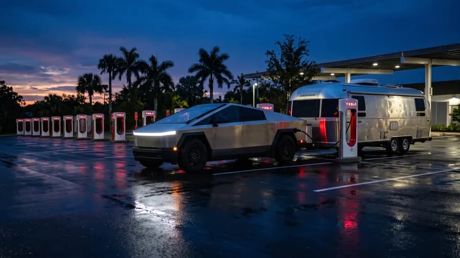 A Cybertruck towing a silver travel trailer, plugged in at a Tesla Supercharger at dusk with the trailer still attached